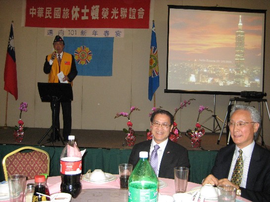 榮光會張連佑會長(左立者)在[邁向101新年春宴]上致歡迎詞。駐處廖處長東周(中)與中華公所共同主席陸忠(右)合影